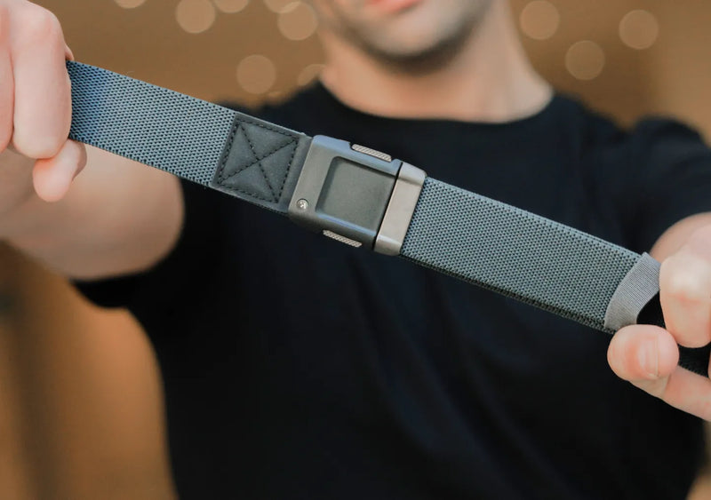 A person wearing a black t-shirt holds out a textured, gray strap with a rectangular, metallic buckle toward the camera, with a blurred background of warm lights.