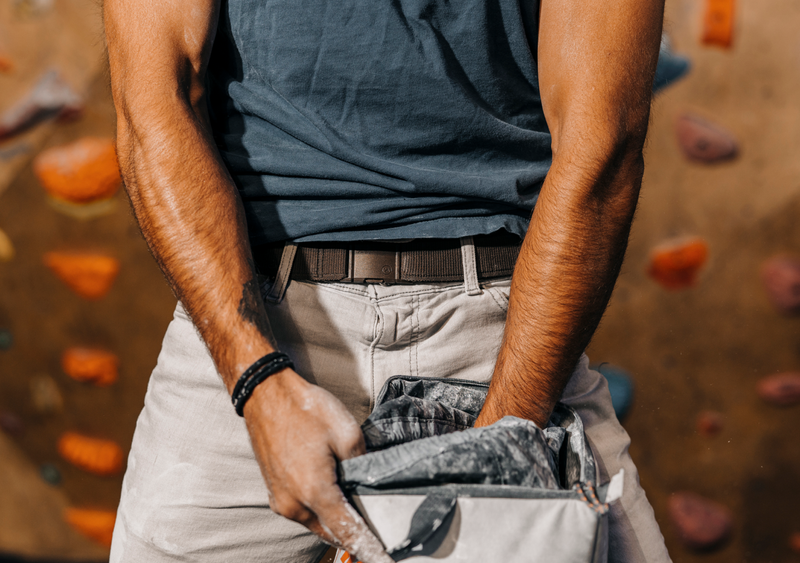 A person in a blue shirt and light pants, secured with a low profile belt, rubs chalk on their hands from a chalk bag, likely preparing to climb, with colorful climbing holds blurred in the background.