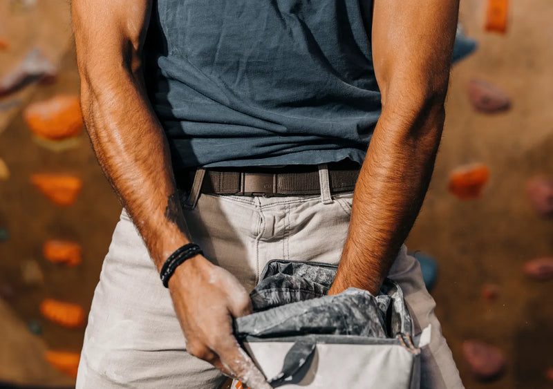 A person wearing a dark t-shirt and light pants dips their hands into a chalk bag, with colorful climbing holds visible in the blurred background.