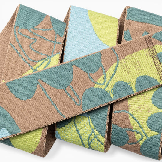 A close-up of the Arcade Belts Inkstorm, featuring woven fabric with a camouflage pattern in green, brown, blue, and yellow. The word ARCADE is stitched into the material, and its performance stretch webbing ensures flexibility for a movement-ready design.