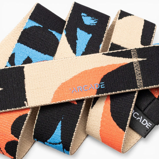Close-up of the Arcade Belts Inkstorm, a woven fabric belt showcasing abstract black, orange, and blue patterns with “ARCADE” stitched in light blue on the beige section. Made with performance stretch webbing for a movement-ready design, this belt combines style and flexibility.