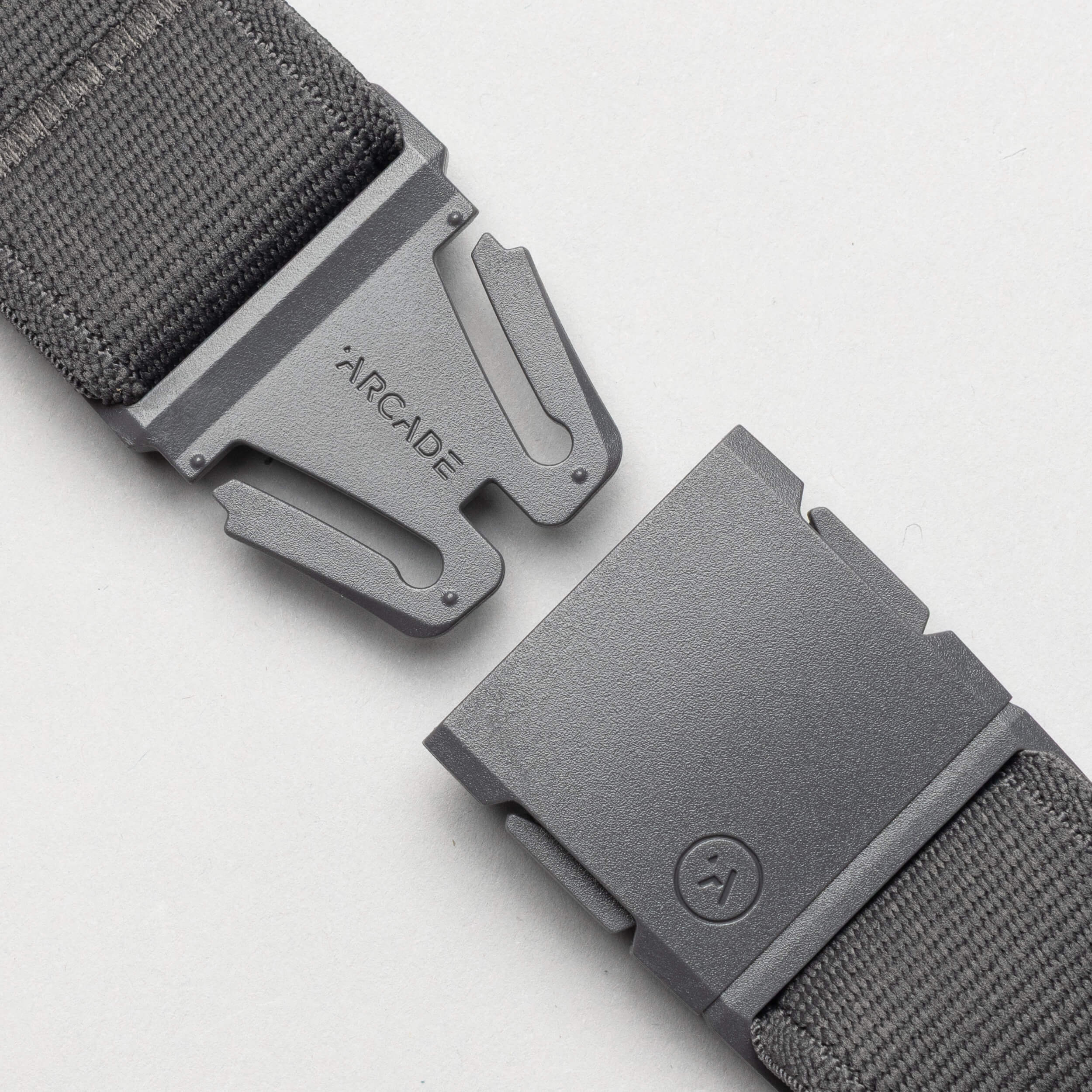 A close-up of an unbuckled Arcade Belts Keep Tahoe Blue gray fabric belt with a metal buckle, one side engraved with "ARCADE" and the other showing a small circular logo, displayed on a light background.