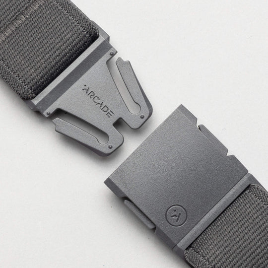 A close-up of an unbuckled Arcade Belts Keep Tahoe Blue gray fabric belt with a metal buckle, one side engraved with "ARCADE" and the other showing a small circular logo, displayed on a light background.