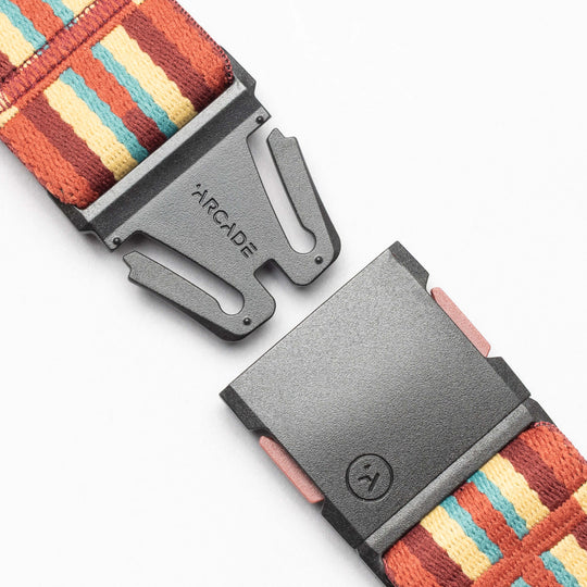 A close-up of a black plastic buckle, engraved with ARCADE, partially unfastened on the Vernan Kee strap by Arcade Belts. The colorful striped fabric features red, yellow, and blue geometric shapes inspired by Navajo rug patterns.