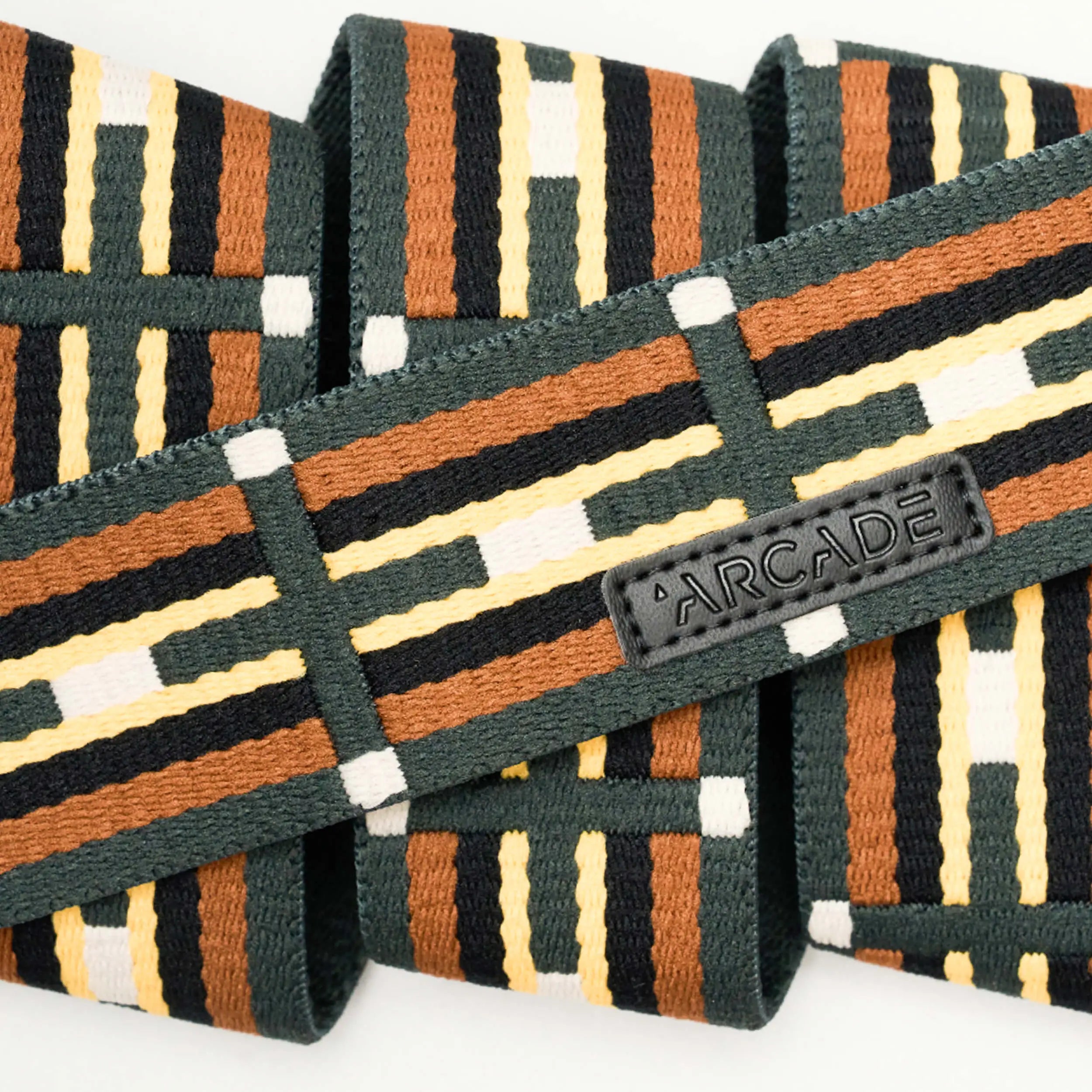 A close-up of the Vernan Kee Elastic Stretch Belt by Arcade Belts, with brown, black, yellow, and white geometric stripes, a stitched black ARCADE label, and flexible stretch webbing.