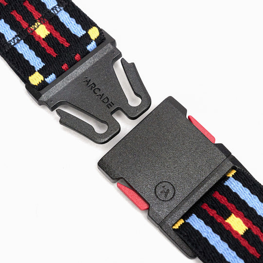 A close-up of a black plastic buckle, with ARCADE printed on it, partially unfastened and attached to a Vernan Kee strap featuring a blue, red, and yellow pattern—showcasing Arcade Belts signature stretch webbing style.