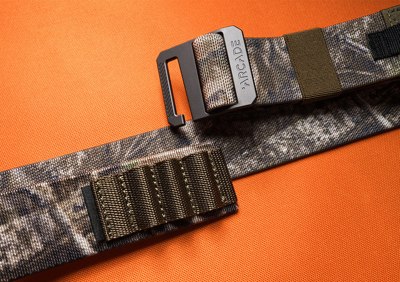 A camouflage-patterned Low Profile Belt by Arcade Belts, featuring stretch webbing and a black metal buckle labeled ARCADE, is displayed on an orange textured surface. The belt boasts sturdy stitching and reinforced sections.