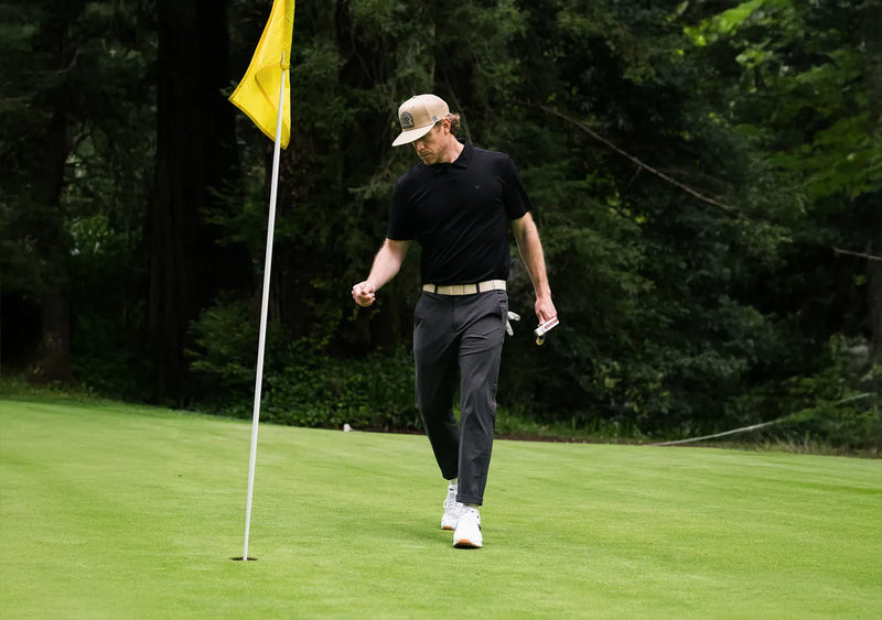 A golfer wearing a black shirt, gray pants, and a beige cap walks near the hole on a green, fist-pumping in celebration. A yellow flag marks the hole, and trees are visible in the background.