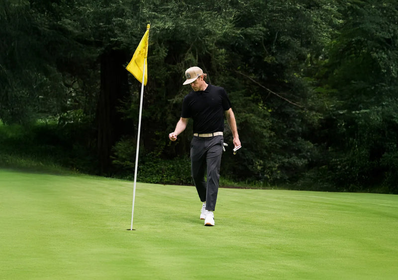 A person wearing a beige cap, black shirt, and gray pants walks on a golf green, holding a golf ball and scorecard near a hole with a yellow flag. Trees and greenery are visible in the background.