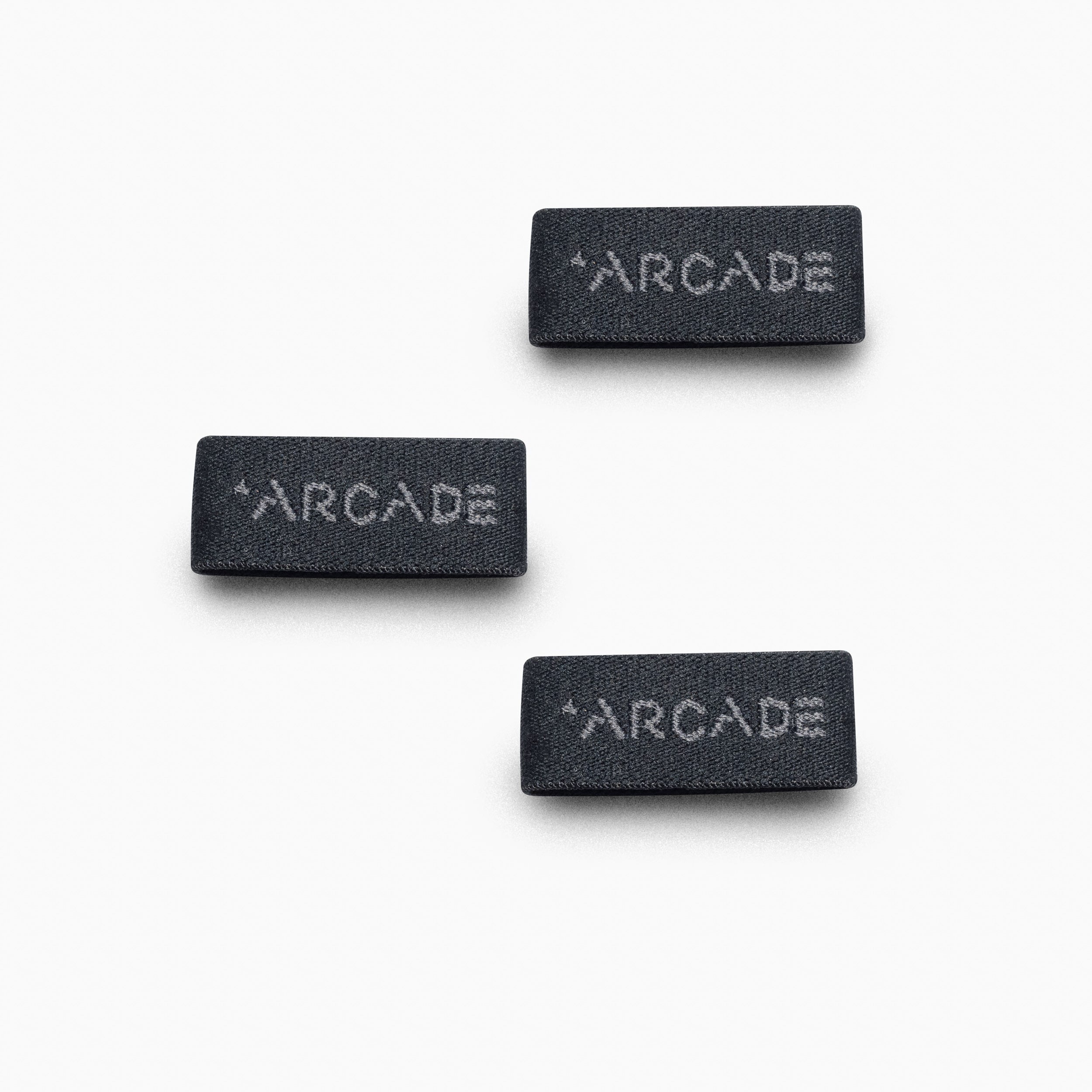 Three rectangular black fabric labels with ARCADE in white are shown on a white background. These scattered Belt Loopkeepers showcase Arcade Belts iconic branding, recognized for innovative Elastic Stretch Belt designs.
