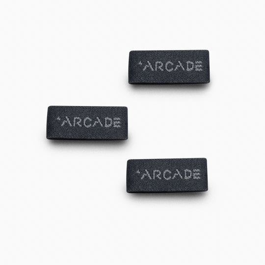Three rectangular black fabric labels with ARCADE in white are shown on a white background. These scattered Belt Loopkeepers showcase Arcade Belts iconic branding, recognized for innovative Elastic Stretch Belt designs.