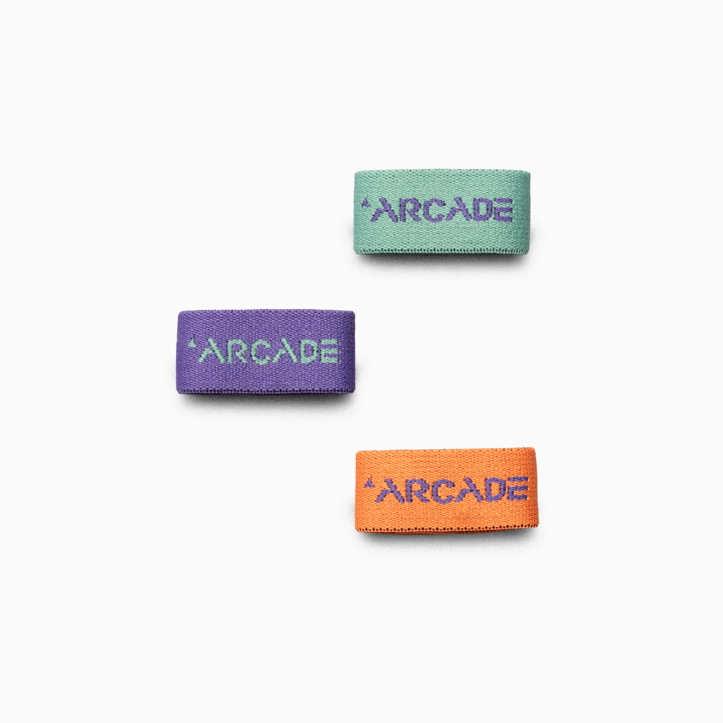 Three Belt Loopkeepers from Arcade Belts—green with purple text, purple with teal text, and orange with purple text reading ARCADE—showcase the brands colorful style and signature Stretch Webbing on a white background.