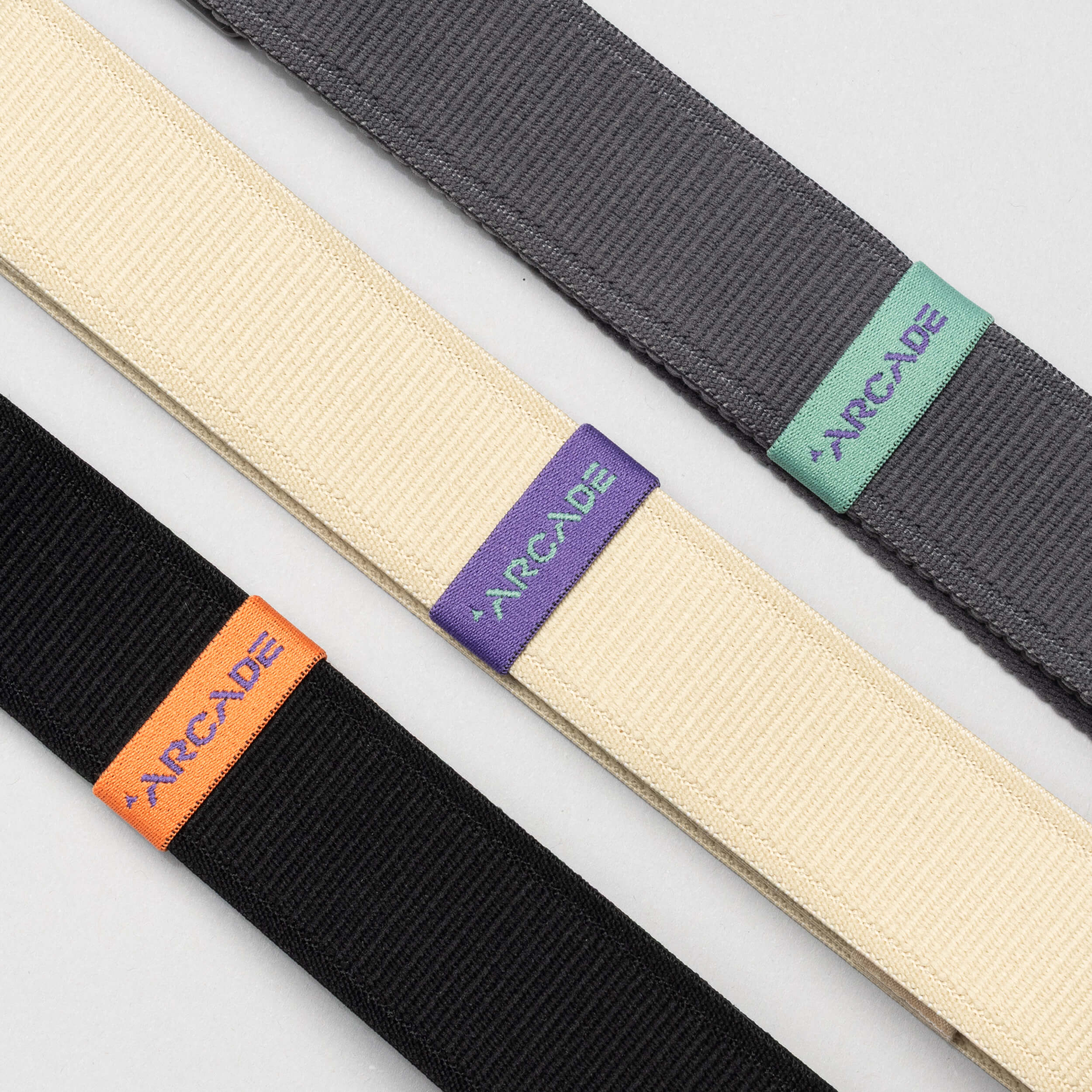 Three Arcade Belts Loopkeepers in black, cream, and gray lie diagonally on a light surface. Each elastic stretch belt features a colored tag with ARCADE in orange, purple, or turquoise, and a sleek plastic buckle.