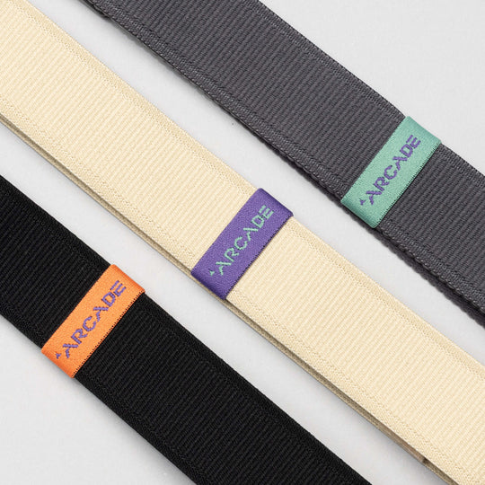 Three Arcade Belts Loopkeepers in black, cream, and gray lie diagonally on a light surface. Each elastic stretch belt features a colored tag with ARCADE in orange, purple, or turquoise, and a sleek plastic buckle.
