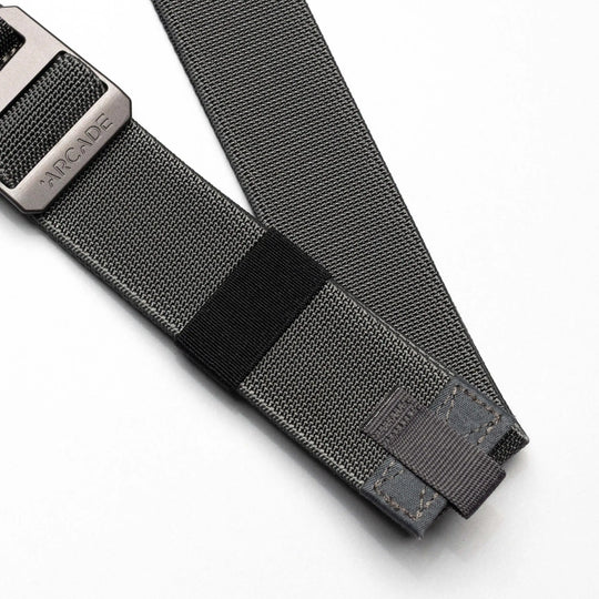 A close-up of the Arcade Belts Hardware Belt Loopkeepers: a grey stretch webbing belt with reinforced stitching and a black metal buckle labeled ARCADE, shown on a white background—ideal for versatile travel wear.