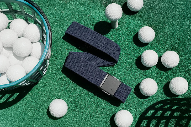 A black textured belt is laid on green turf surrounded by white golf balls, some in baskets and others scattered around, with a single ball on a tee.
