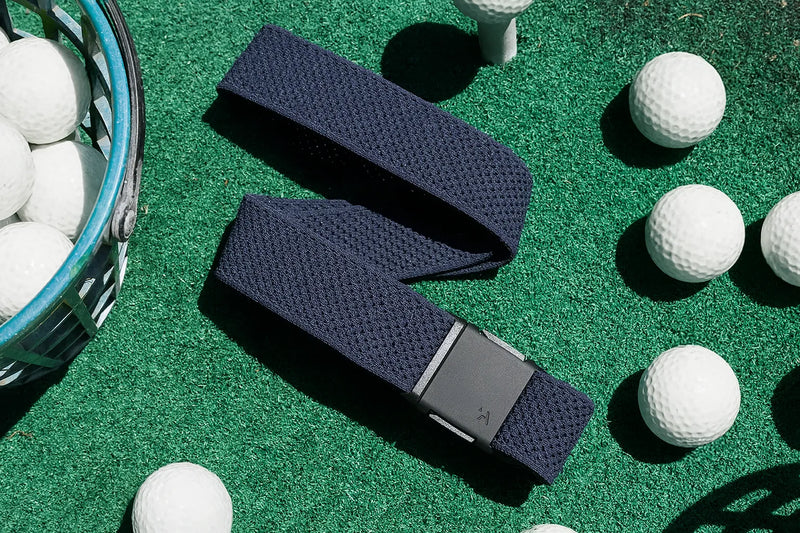 A navy blue woven belt with a black buckle is placed on green turf surrounded by several white golf balls and part of a wire basket.