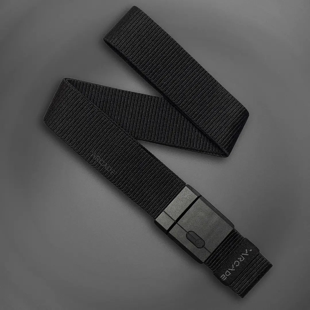 A black tactical nylon belt with a rectangular black buckle is laid out on a flat, dark gray surface. The belt features the word ARCADÉ printed near the end.
