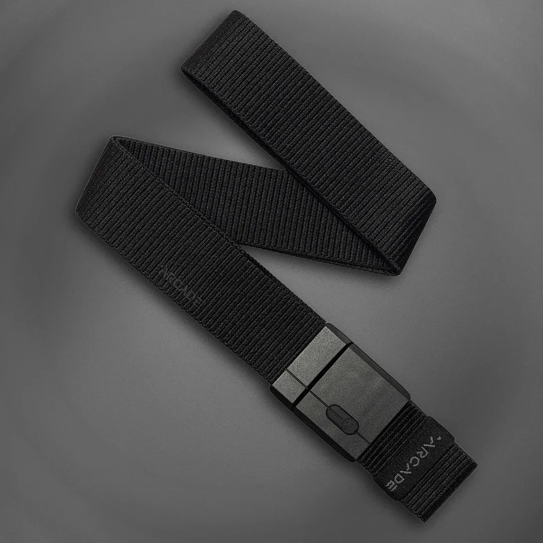 A black nylon belt with a matte black rectangular buckle is laid out on a gray background. The belt has a textured surface and ARCANE branding near the end.