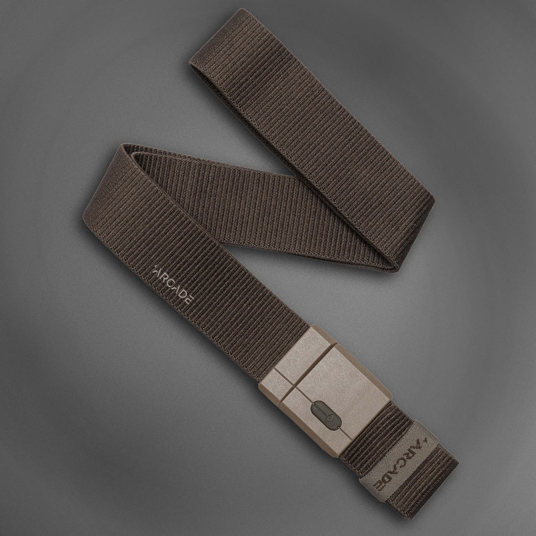 A brown, woven fabric belt with a rectangular buckle, displayed on a gray background. The belt is neatly coiled with the brand name ARCADE visible on the end.