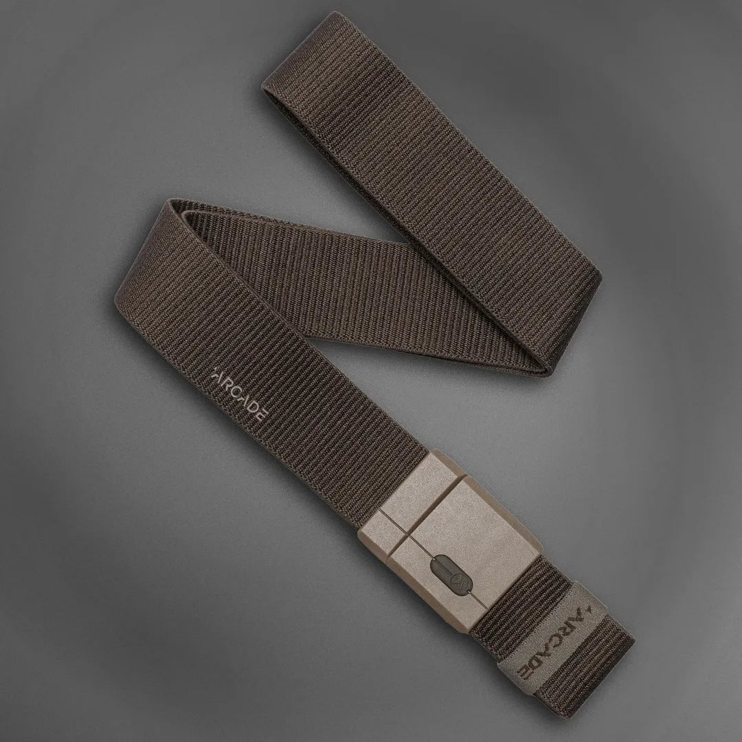 A brown, adjustable nylon belt with a rectangular plastic buckle is displayed on a gray background. The buckle has a simple clasp mechanism and the word ARCADER is visible near one end of the belt.