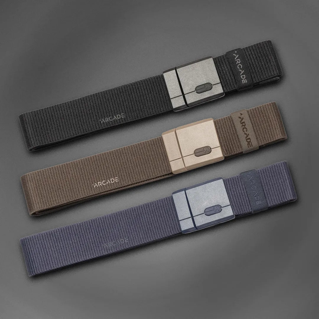 Three fabric belts in black, brown, and navy are laid out diagonally on a gray background. Each belt has a rectangular buckle and the brand name ARCADE printed near the buckle.