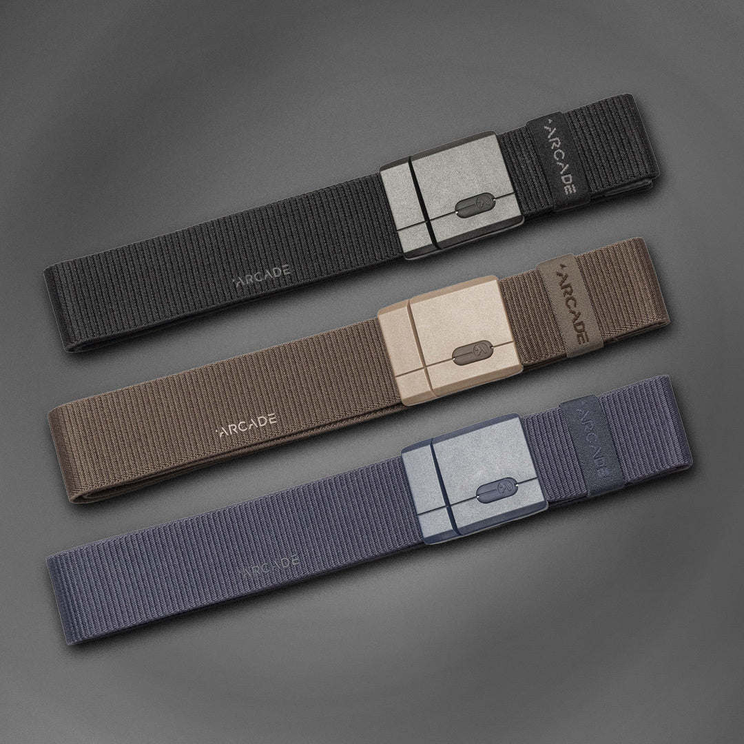 Three fabric belts in black, brown, and navy are laid flat on a gray surface. Each belt has a matching rectangular buckle and the word ARCADE printed near the buckle.