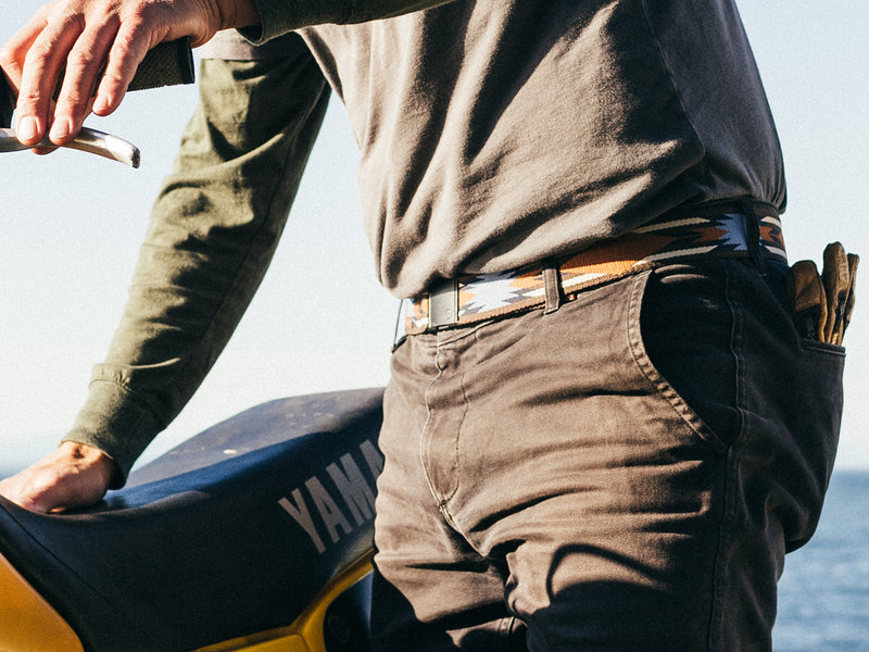 A person wearing dark pants, an Arcade Belt with a plastic buckle, and a green long-sleeve shirt stands next to a motorcycle seat labeled YAMAHA. They hold sunglasses and have tools tucked in their back pocket.