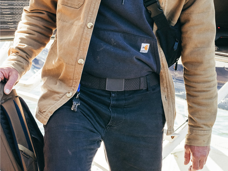 A person wearing a tan jacket, blue hoodie, and black jeans holds a backpack in one hand and keys in the other. A small black bag with a plastic buckle is strapped across their chest.