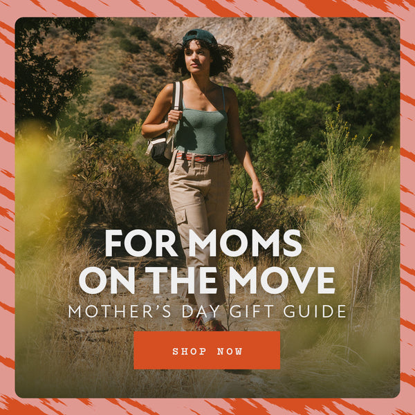 A woman hikes outdoors carrying a backpack, surrounded by greenery and hills. Text overlay reads: For Moms On The Move. Mother’s Day Gift Guide featuring Arcade Belts. A button below says Shop Now. Orange brushstroke border frames the image.