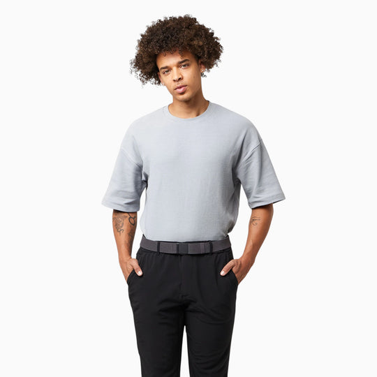 Wearing the Arcade Belts Momentum Kit, a person with curly hair stands against a white background. Their loose gray short-sleeve shirt is tucked into black pants and cinched with the sleek Momentum belt—an effortlessly stylish choice from Arcade Belts that’s sure to become a wardrobe staple. With hands in pockets and a calm, neutral expression, they embody relaxed confidence and everyday comfort.