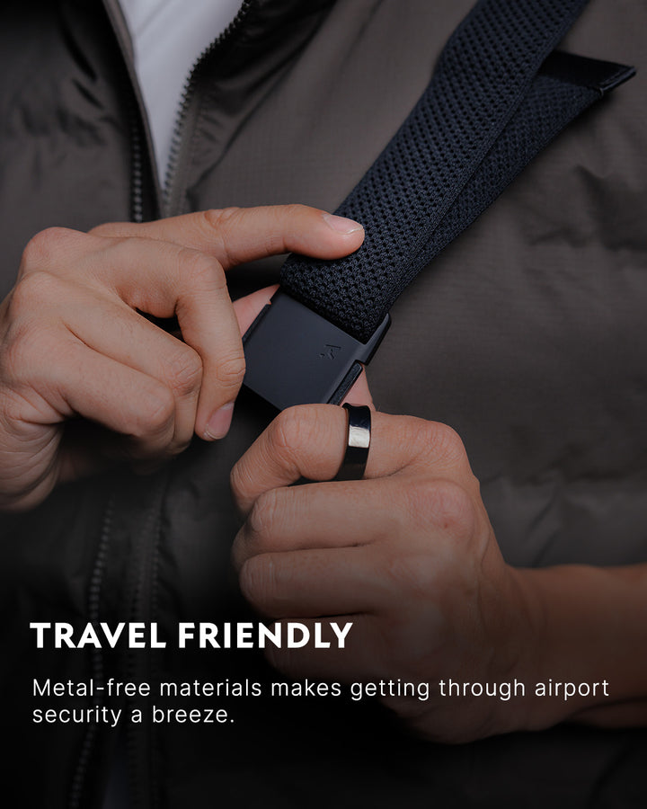Close-up of a person adjusting a low profile belt with a plastic buckle, wearing a dark jacket and a ring. Text reads: TRAVEL FRIENDLY. Metal-free materials make travel belts ideal for breezing through airport security.