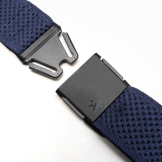 Close-up of the Arcade Belts Momentum stretch webbing strap in navy blue, featuring a black plastic clasp thats partly unfastened, shown on a light gray background.