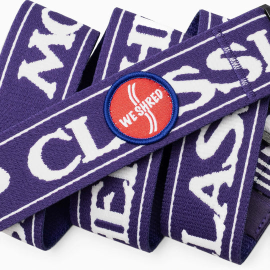 A close-up of a purple Arcade Belts fabric band featuring bold white text and a round red patch outlined in blue that reads “WE SHRED” in white, designed to commemorate the Mothership Classic—a signature event supporting the High Fives Foundation.