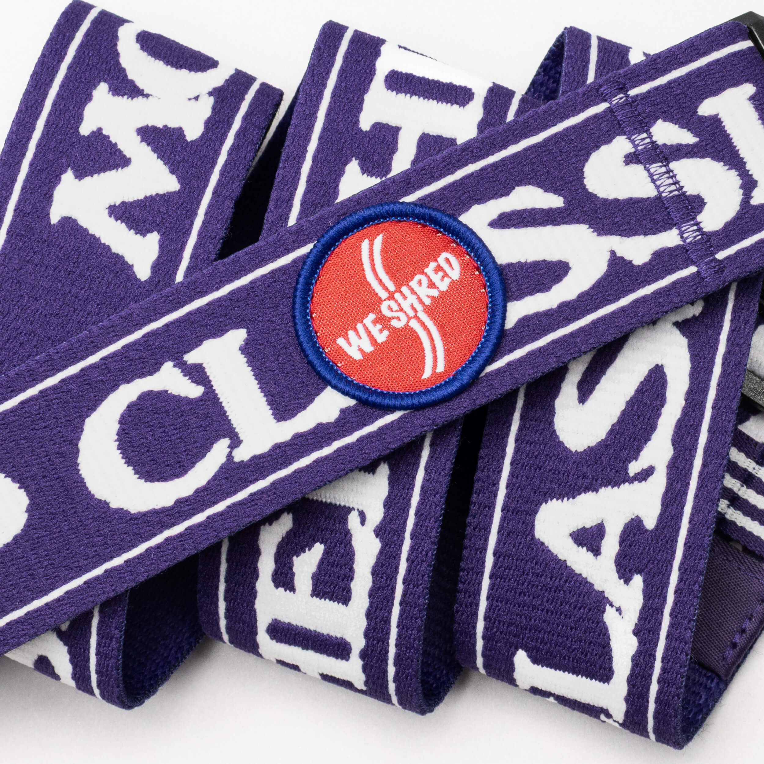 The Mothership Classic by Arcade Belts features a purple strap with bold white text and a round red patch reading "WE SHRED" in white, outlined in blue. Inspired by the KT-22 chair lift, it's displayed in overlapping loops on a white background.