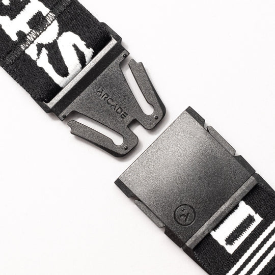 Close-up of the Arcade Belts Mothership Classic, featuring stretch webbing and a low profile. The black plastic buckle is partially unclipped, showing ARCADE on the metal piece. The strap displays white text and patterns.