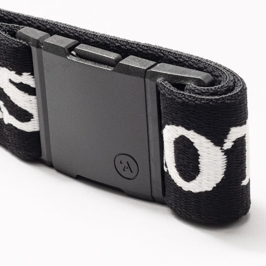 Close-up of Arcade Belts Mothership Classic: black stretch webbing strap with white embroidered letters and a rectangular plastic buckle, shown on a white background.