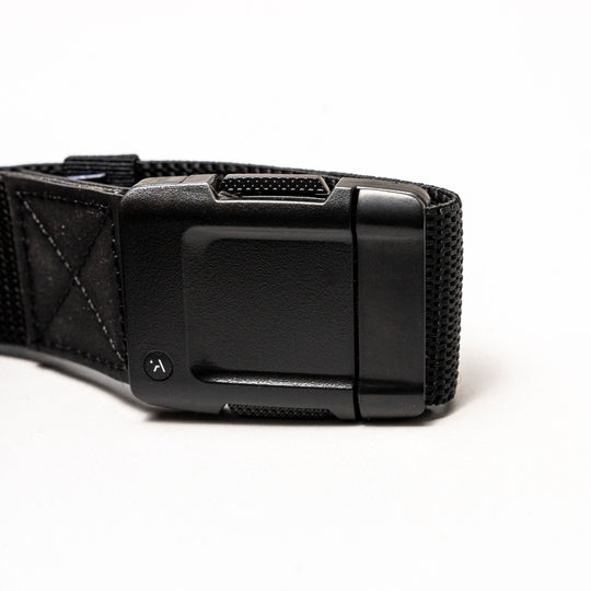 Close-up of the Arcade Belts Motion: a black fabric strap with a rectangular, textured plastic buckle on a white background. The buckle has a small round button with a curved arrow for secure fastening and easy adjustment—perfect for travel.