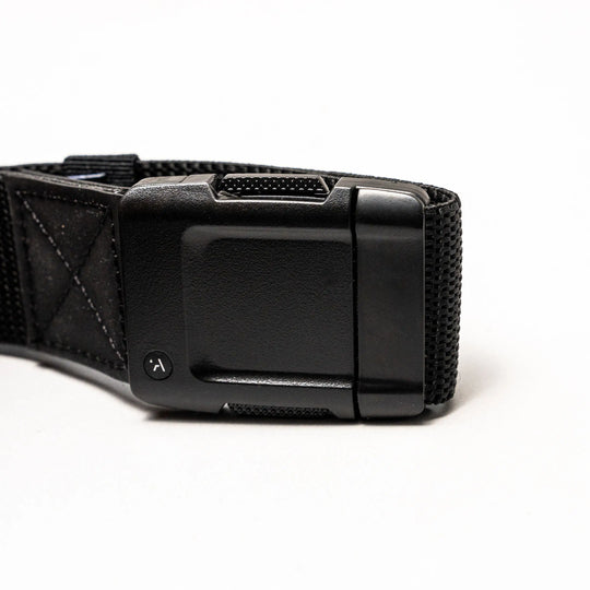 Close-up of the Arcade Belts Motion: a black fabric strap with a rectangular, textured plastic buckle on a white background. The buckle has a small round button with a curved arrow for secure fastening and easy adjustment—perfect for travel.