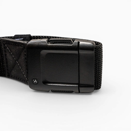 Close-up of the Arcade Belts Motion: a black fabric strap with a rectangular, textured plastic buckle on a white background. The buckle has a small round button with a curved arrow for secure fastening and easy adjustment—perfect for travel.