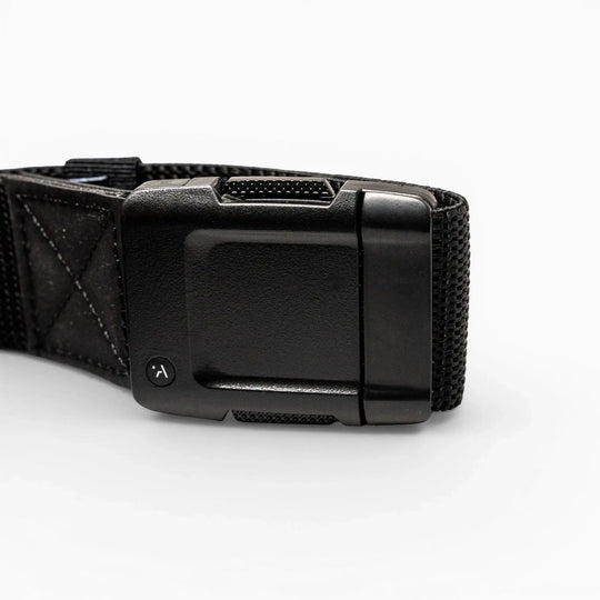 Close-up of the Arcade Belts Motion: a black fabric strap with a rectangular, textured plastic buckle on a white background. The buckle has a small round button with a curved arrow for secure fastening and easy adjustment—perfect for travel.