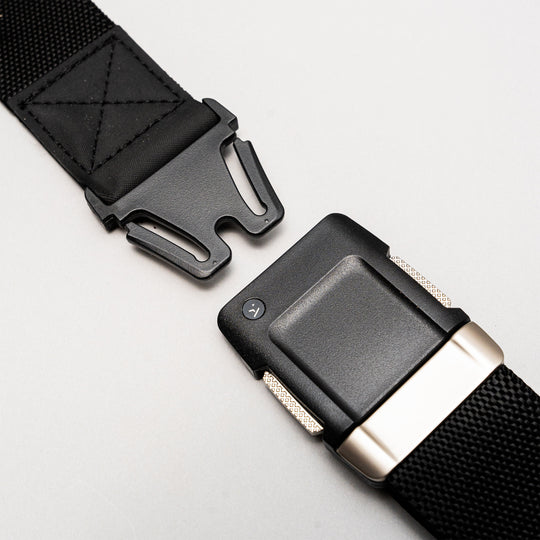 Close-up of the Arcade Belts Motion: a black nylon belt with a sturdy, quick-release black plastic buckle featuring metallic accents. The partially unfastened buckle highlights its clean, modern design against a light gray background.