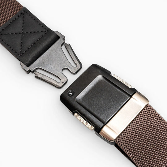 Close-up of the Arcade Belts Motion brown woven low-profile belt with a black and silver buckle and leather detail near the buckle, shown partially unfastened on a light grey background.