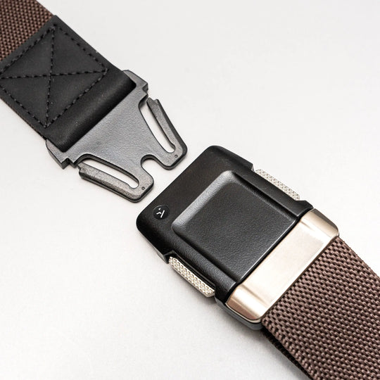 Close-up of the Arcade Belts Motion brown woven low-profile belt with a black and silver buckle and leather detail near the buckle, shown partially unfastened on a light grey background.