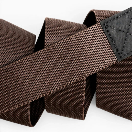 Close-up of the Arcade Belts Motion belt, featuring brown textured nylon stretch webbing and a black stitched patch, coiled loosely on a white background.