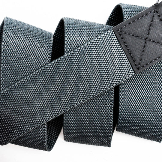 Close-up of a black, textured nylon strap from the Motion by Arcade Belts, looped in a zigzag pattern like a Low Profile Belt, featuring a stitched rectangular black leather patch at one end on a white background.