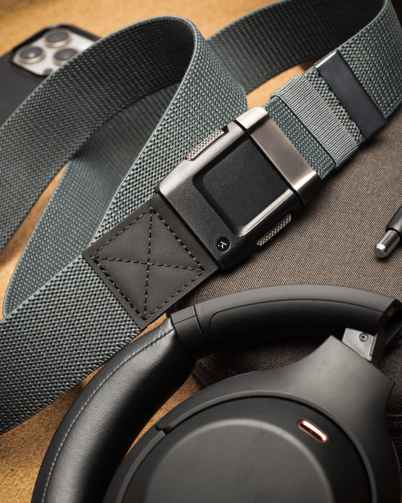 A close-up of a grey Arcade Belt with a low profile metal buckle, black over-ear headphones, part of a pen, and the corner of a smartphone on a brown surface.