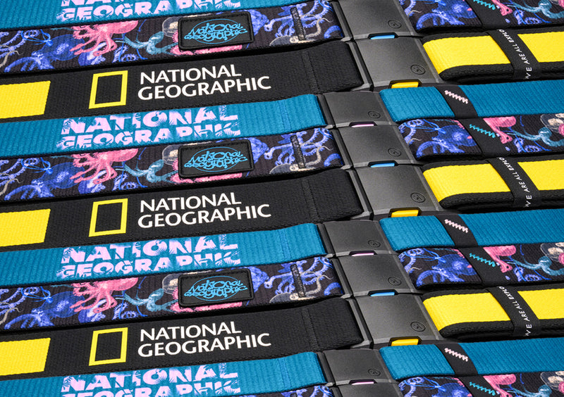Several patterned fabric belts are arranged in rows. They feature the National Geographic logo, a yellow rectangle beside white text, and vibrant designs in blue, yellow, and purple with black plastic buckles.