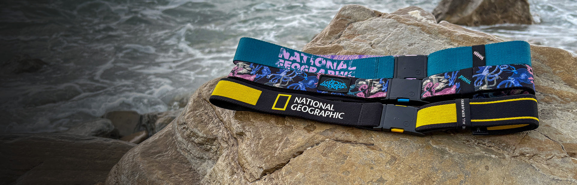 Two colorful National Geographic branded belts with plastic buckles lie on large rocks near the ocean, with waves and rocky shore visible in the background.