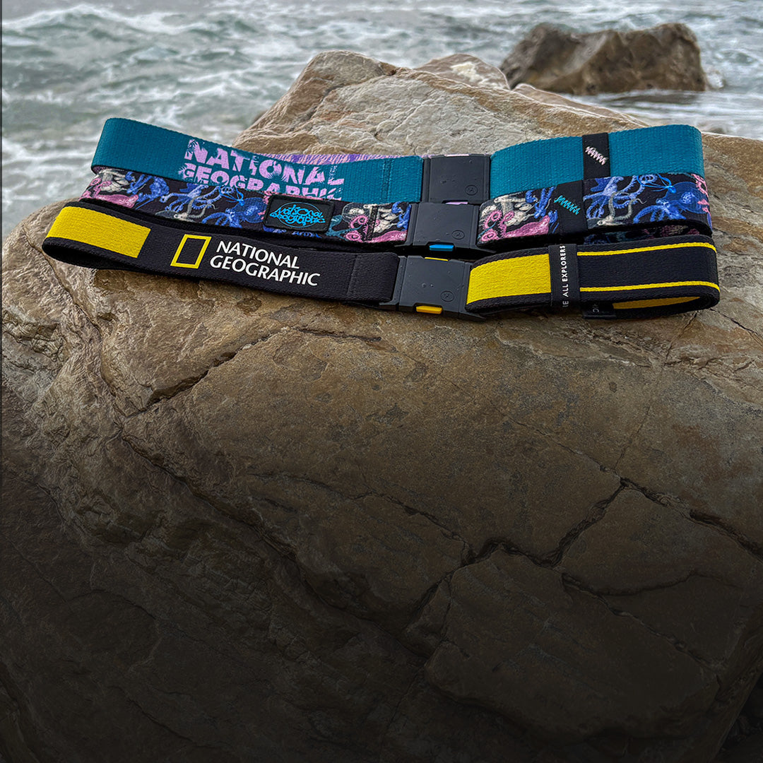 Three National Geographic-branded fabric belts with colorful patterns and black plastic buckles are displayed on a large rock near the ocean, with waves and rocks in the background.